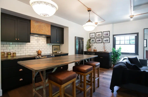 Modern kitchen with black cabinets, white subway tiles, a wooden island, and stylish lighting fixtures. Cozy seating area nearby.